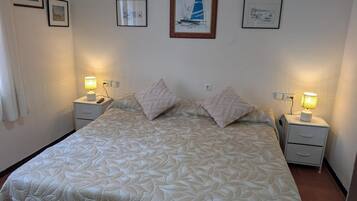 A bedroom with a bed, two bedside tables, a nightstand, and a wall with framed pictures.