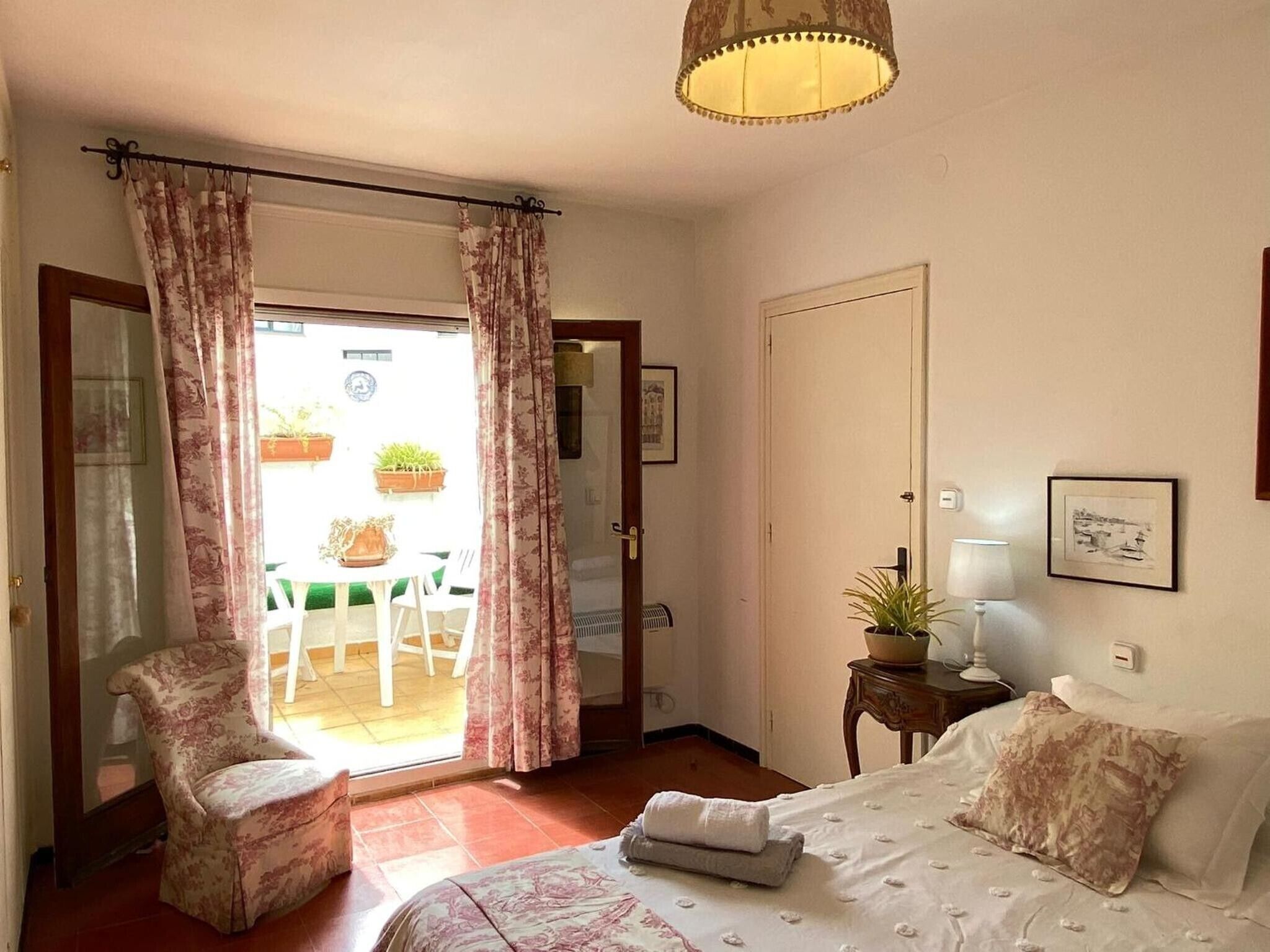 Une chambre à coucher comprenant un lit, une chaise, une table, une lampe et une porte donnant sur un balcon agrémenté de plantes.