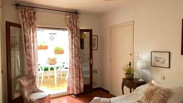 Un dormitorio con cama, una silla, una mesa, una lámpara y una puerta que da a un balcón con plantas.