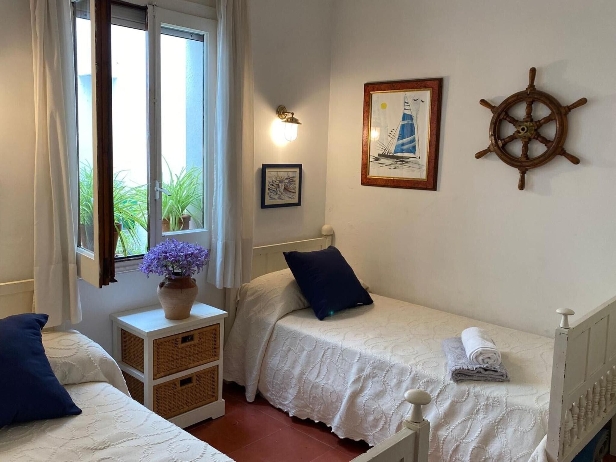 Un dormitorio con dos camas individuales, una ventana con cortinas, una rueda de barco montada en la pared y un cuadro enmarcado.