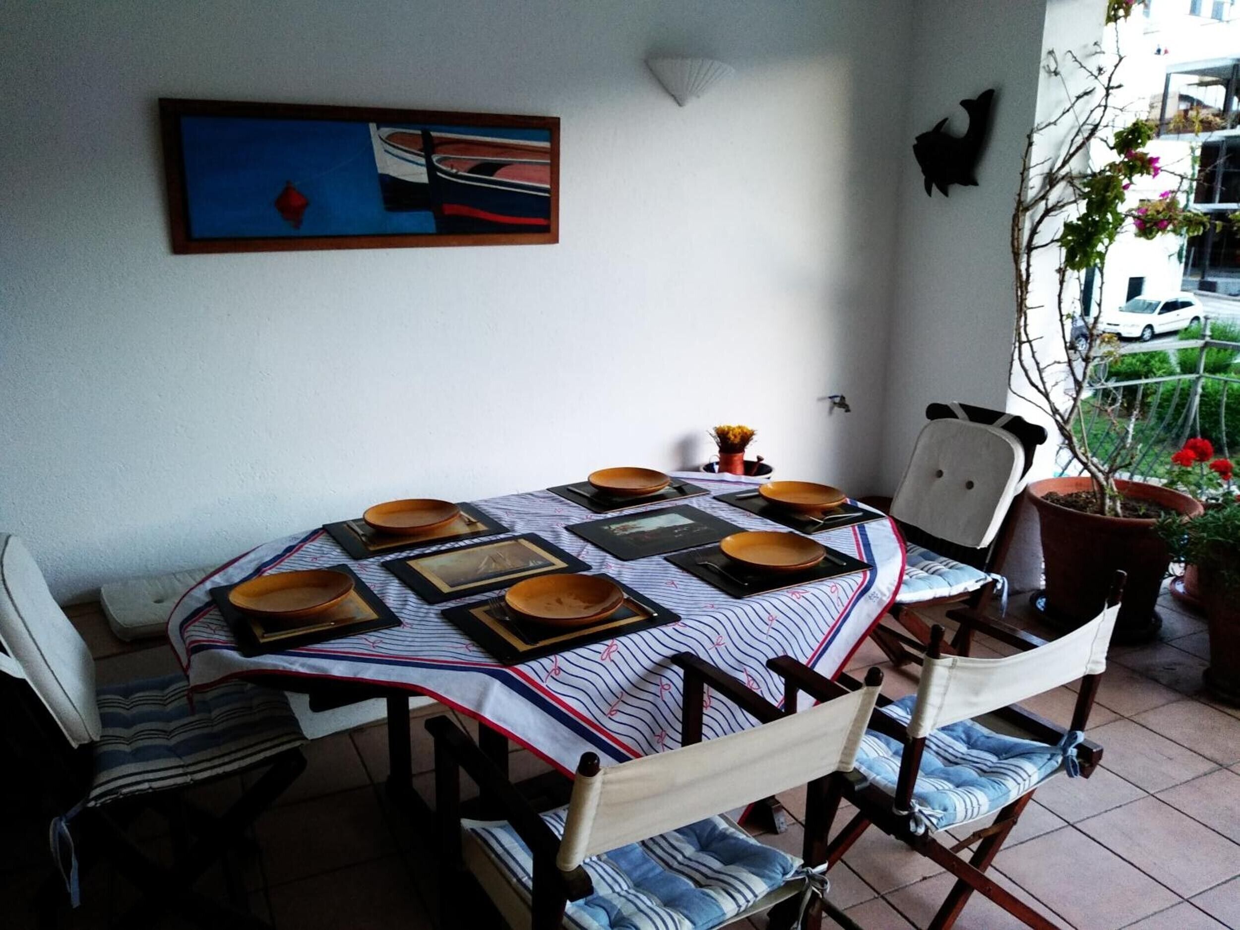 Ein Essbereich mit einem Tisch für vier Personen, Stühle mit gestreiften Kissen, einem gerahmten Bild an der Wand und Topfpflanzen auf dem Balkon.