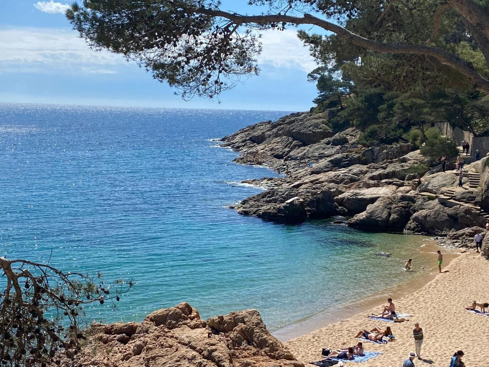En strand med en stenig terräng, kristallklart blått vatten och människor som solar.