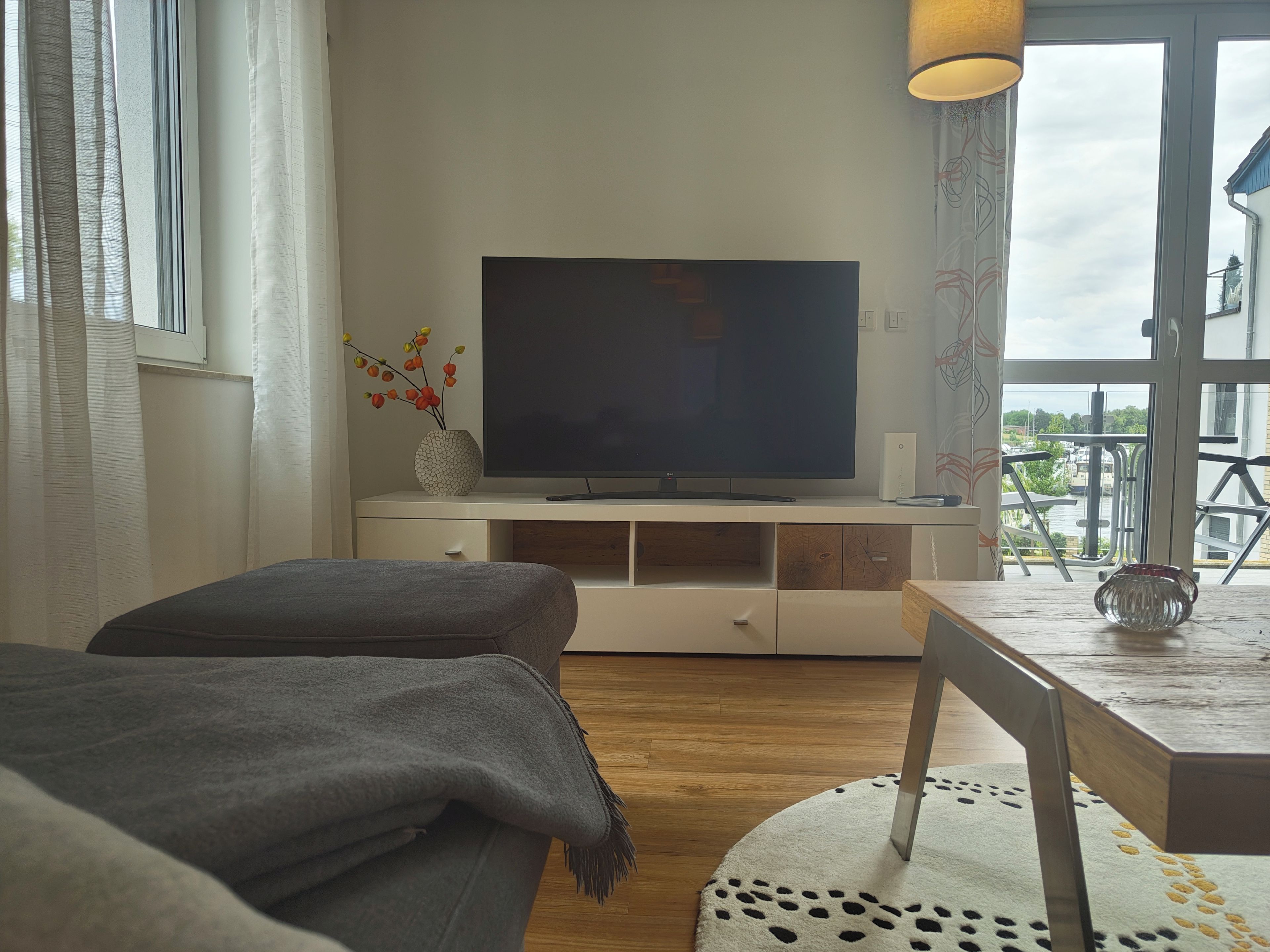 Un salon avec un téléviseur à écran plat posé sur un meuble de télévision blanc, un canapé gris, une petite table avec un vase, et une vue sur un balcon aménagé avec du mobilier d’extérieur.