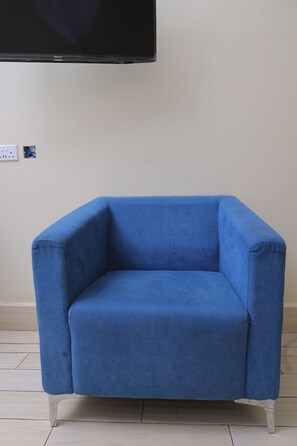 A blue armchair with a straight backrest and a flat seat, positioned against a plain wall with a power outlet and a television mounted above it.