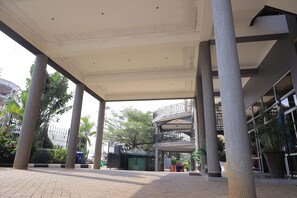 An open-air covered walkway with columns, a multi-level building in the background, and potted plants.