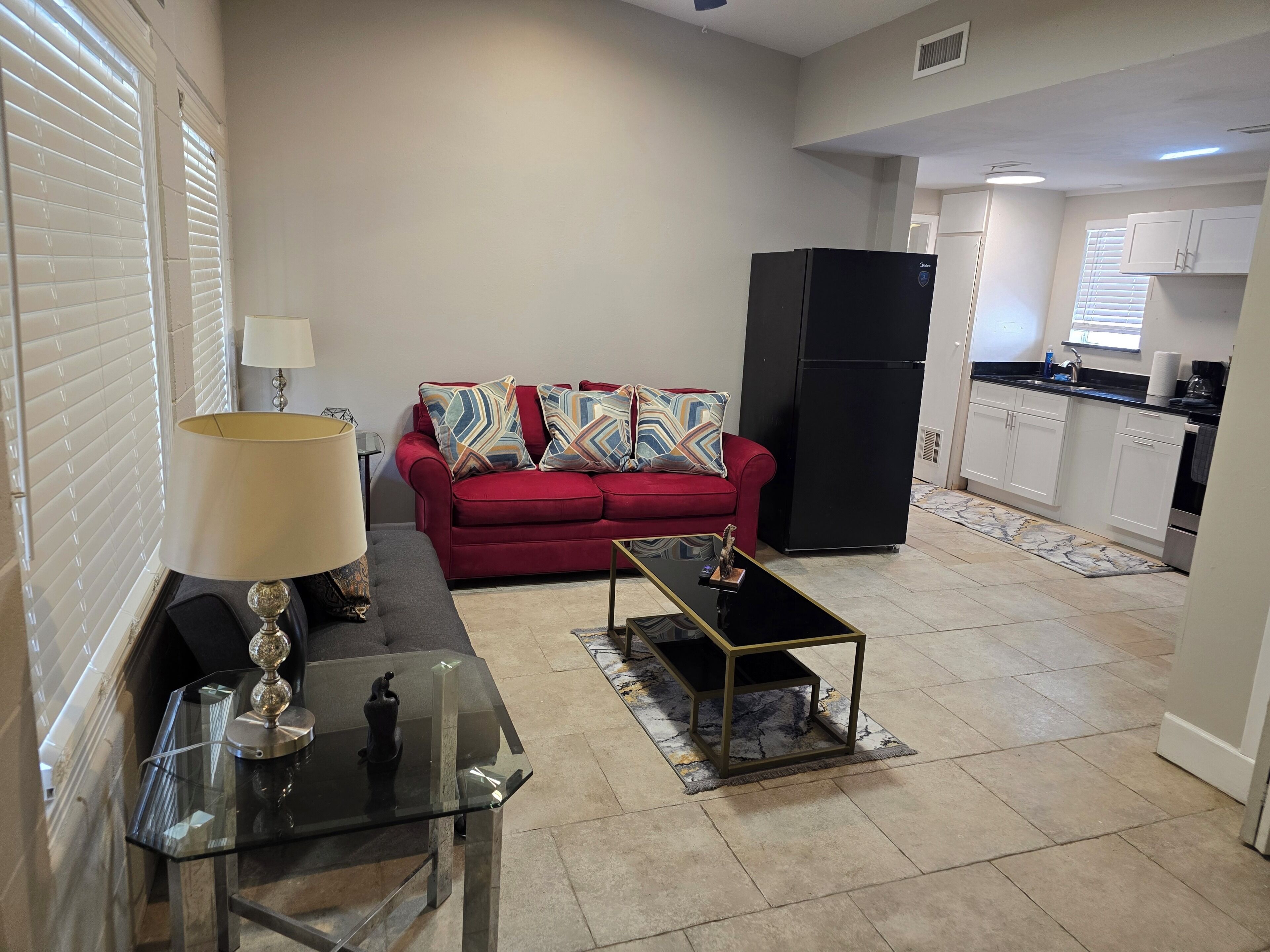 A living room with a red sofa, a glass coffee table, and a lamp. There is a kitchen area with white cabinets and a black refrigerator.