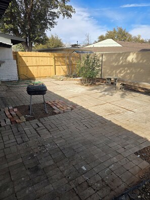 Ein Garten mit Pflasterterrasse, Grill, Holzlattenzaun und einem kleinen Baum.