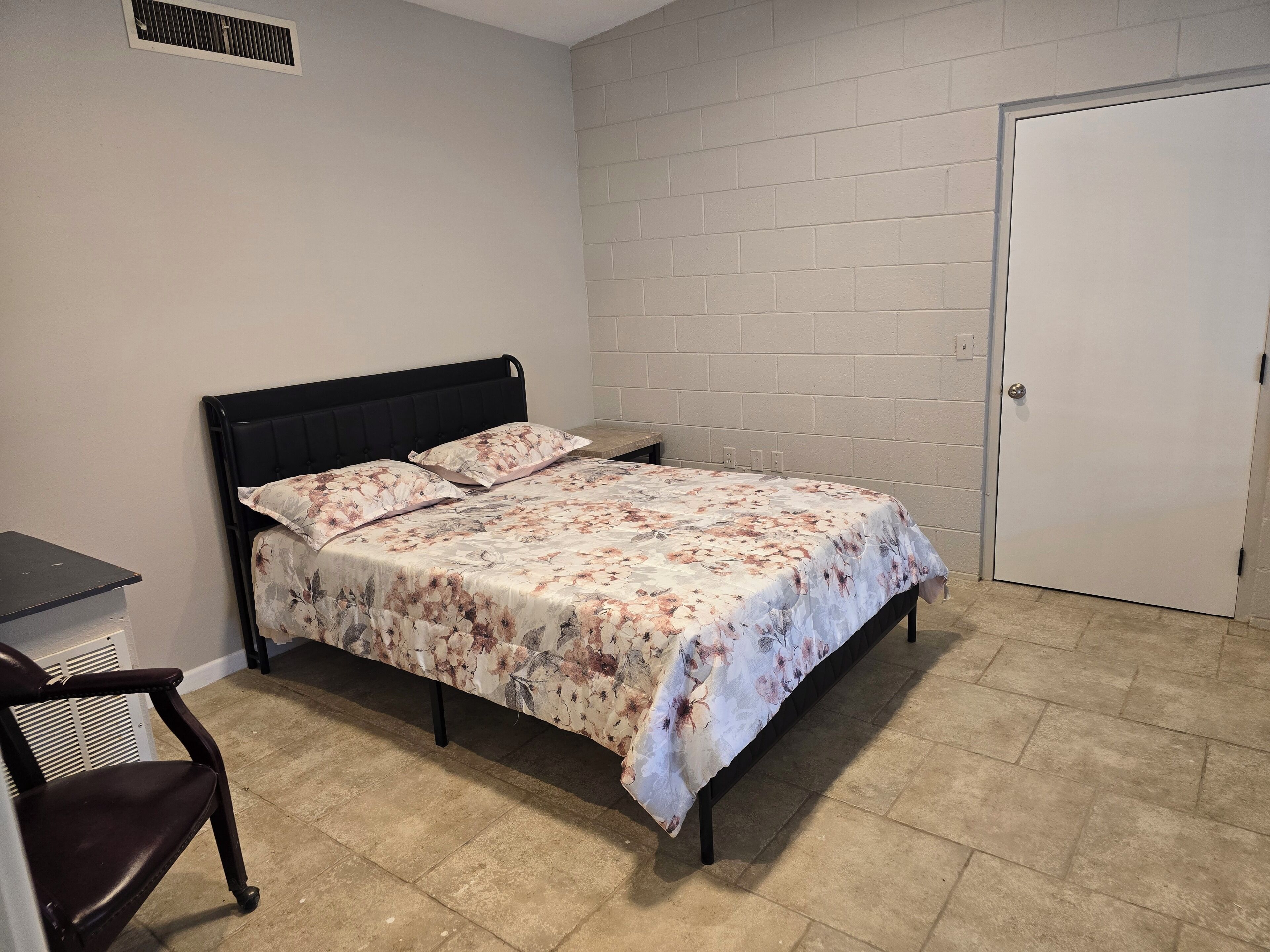 Une chambre avec un lit, une chaise, une porte et un mur muni d’une grille d’aération.