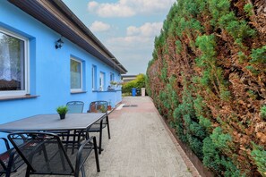 Eine Terrasse mit Tisch und Stühlen, ein blaues Gebäude und ein Hecken.