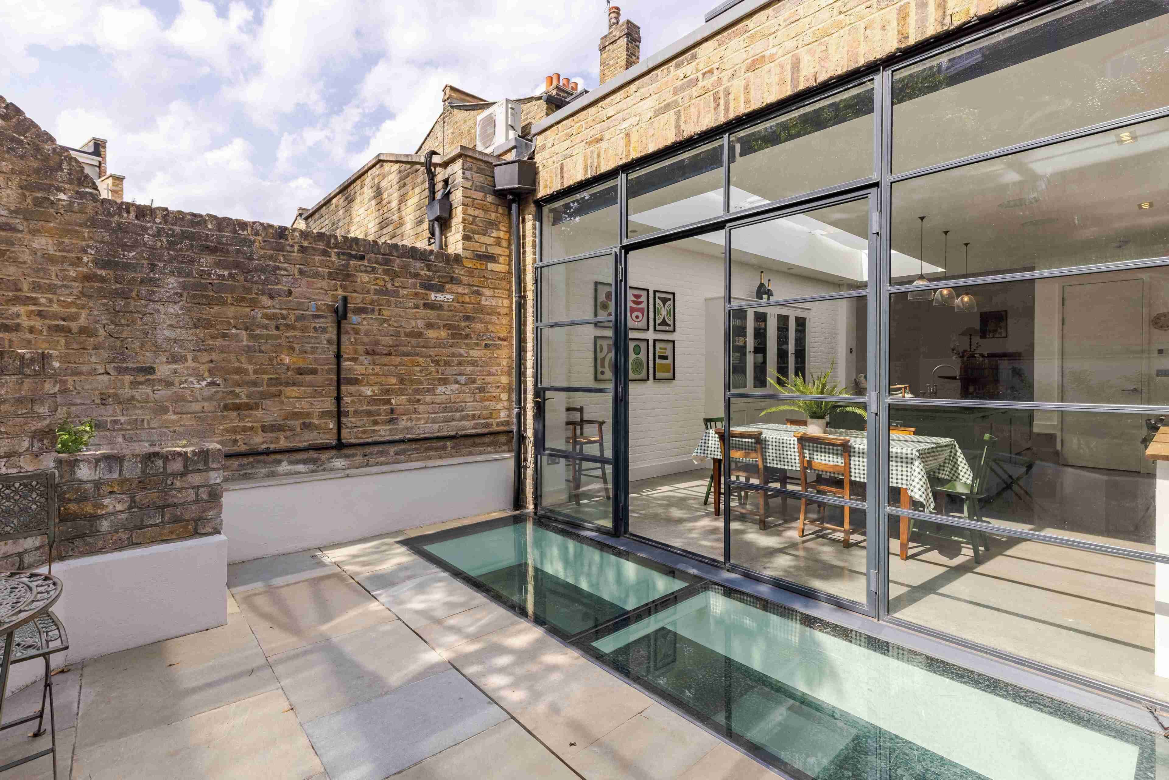 Un patio moderne doté d’une véranda en verre, d’un mur en briques et d’un espace repas avec des tables et des chaises.