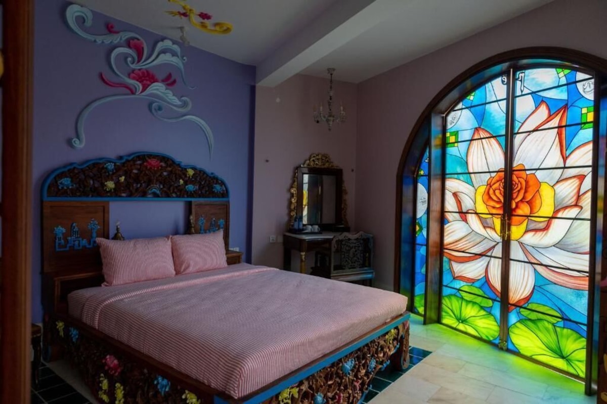 Ein Schlafzimmer mit einem großen Bett, einem verzierten Kopfteil, einem Buntglasfenster und einer Chandelüster.