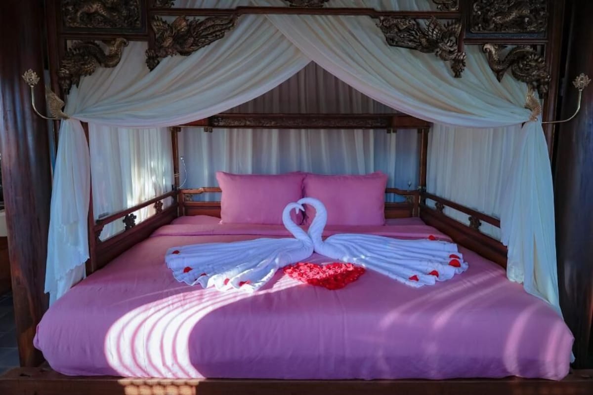 Ein Bett mit Baldachin, einer rosa Tagesdecke und einer zu einem Schwan geformten Handtuchdeko.