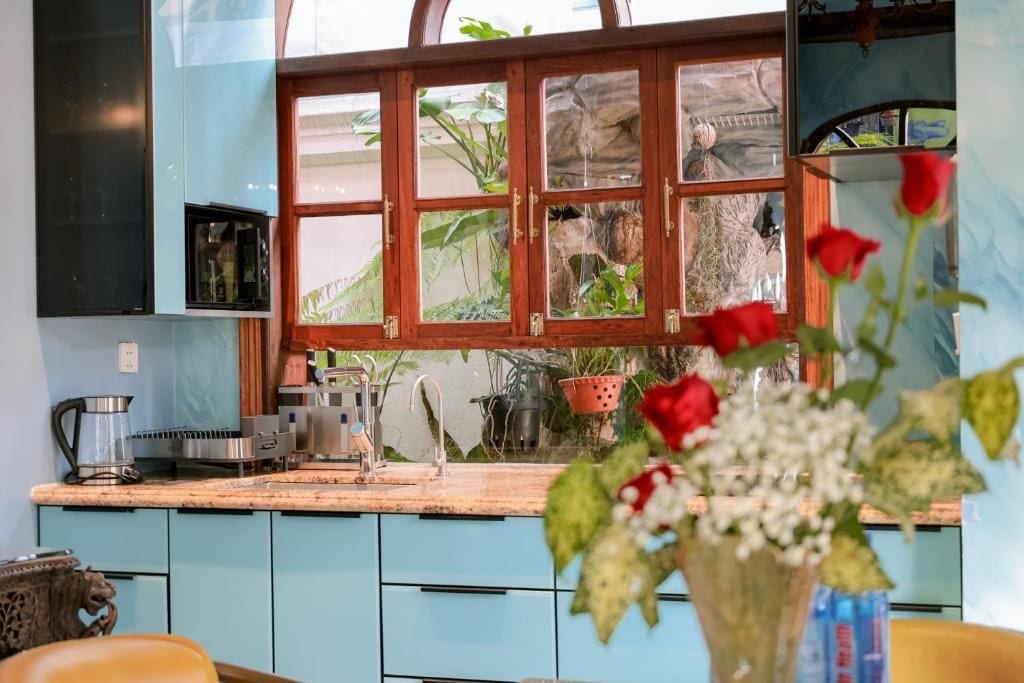 Een keuken met lichtblauwe kastjes, een houten raam met ruitverdeling en een vaas met bloemen op het aanrecht.