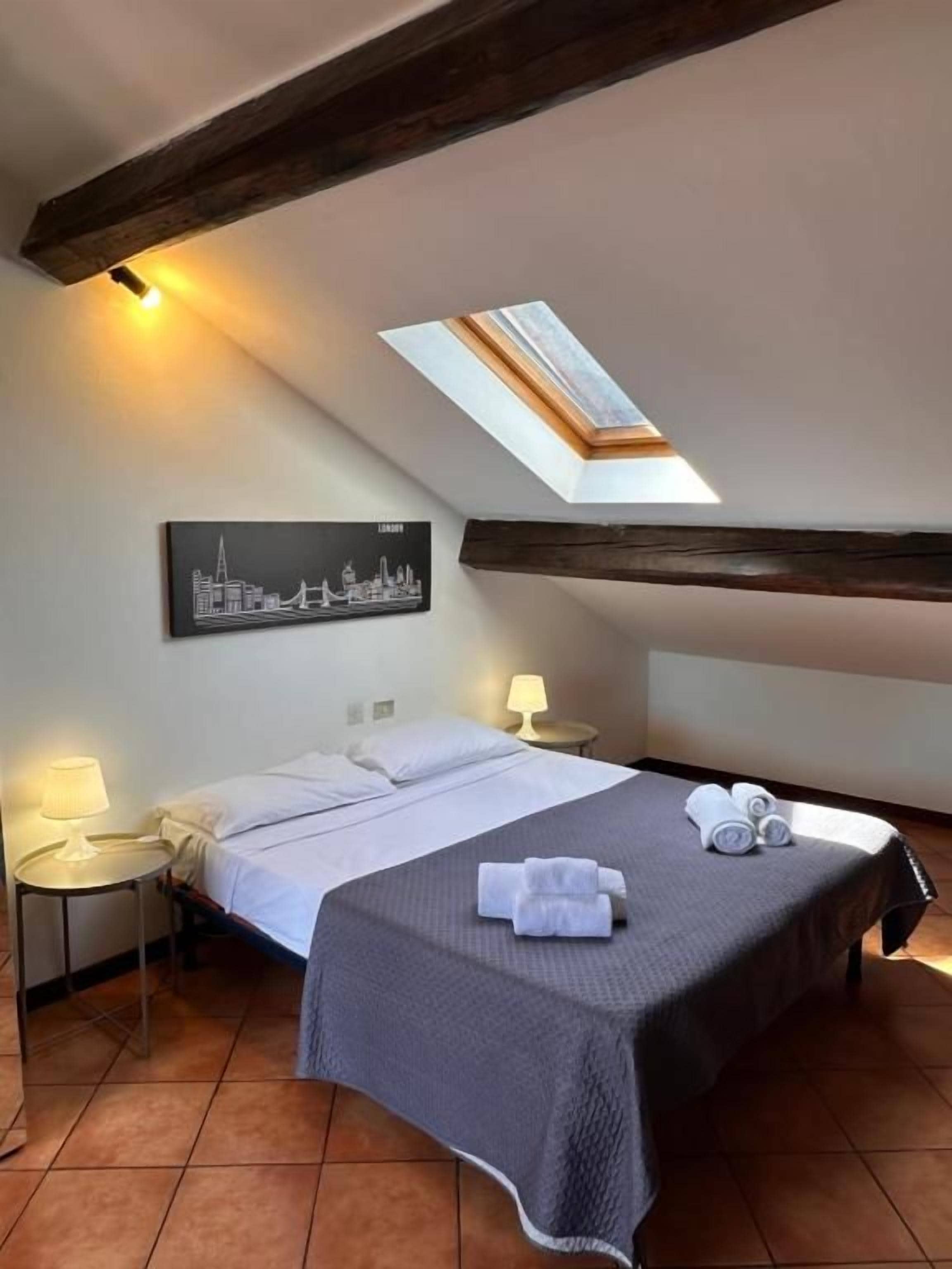 Una habitación de hotel con cama, dos mesitas de noche, un tragaluz y una obra de arte con un paisaje urbano en la pared.