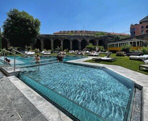 Piscina com água cristalina, rodeada por espreguiçadeiras e um edifício com arcos ao fundo.
