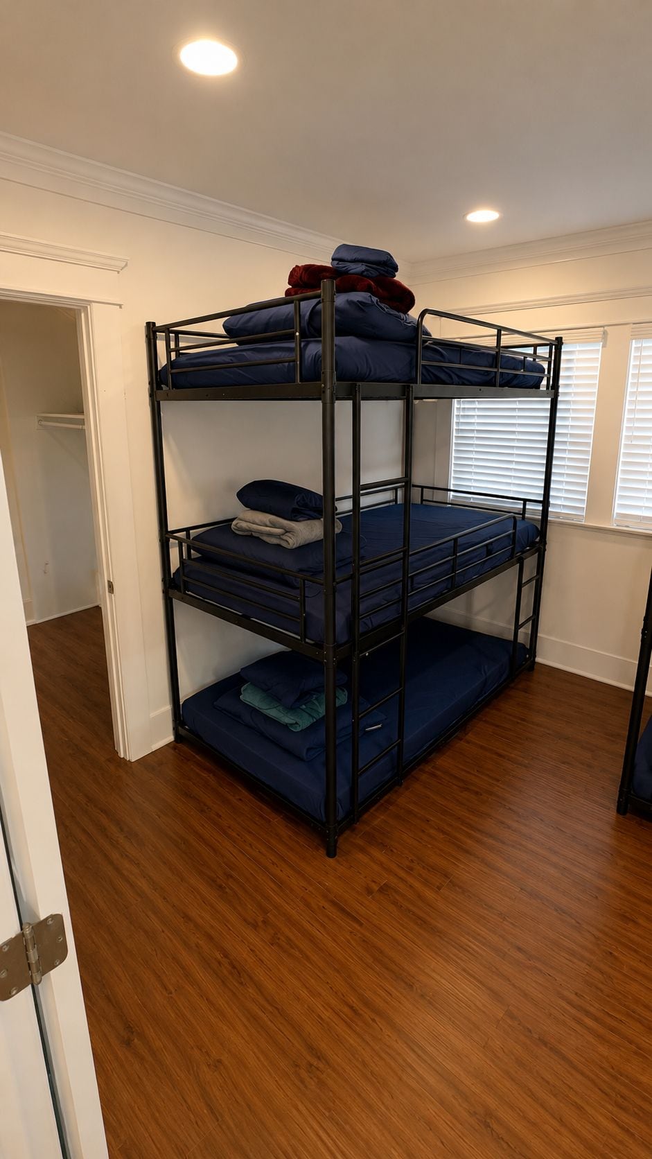 Ein Etagenbett mit drei Schlafstellen, Holzboden und ein Fenster mit Jalousien.