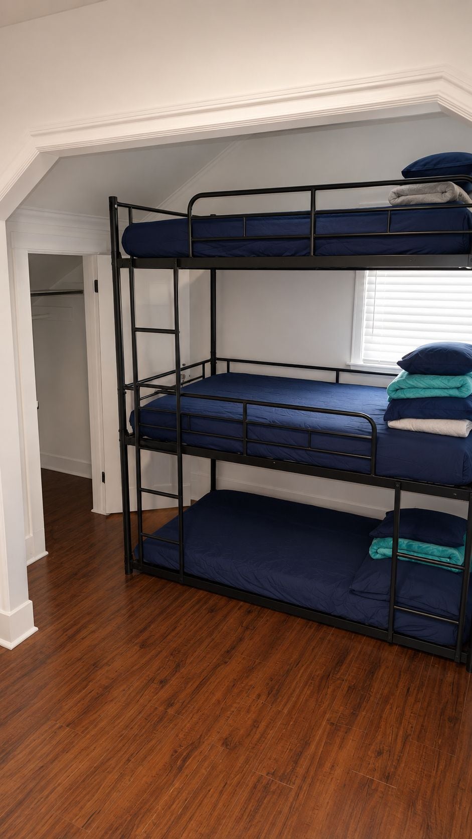 A bunk bed with three levels, a wooden floor, and a window with blinds.