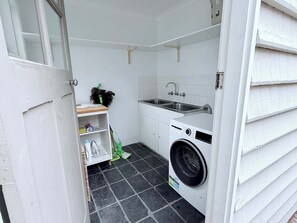 A compact laundry room with a washing machine, a sink, and a small shelf with cleaning supplies. - Harvey’s Beach House Close to South Beach (South Fremantle)