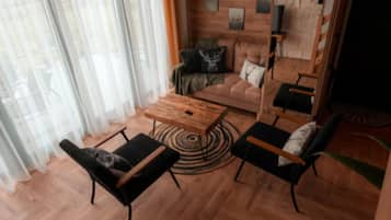 A modern living room with a wooden floor, a round coffee table, two black chairs, a sofa with decorative pillows, and a large window with sheer curtains.