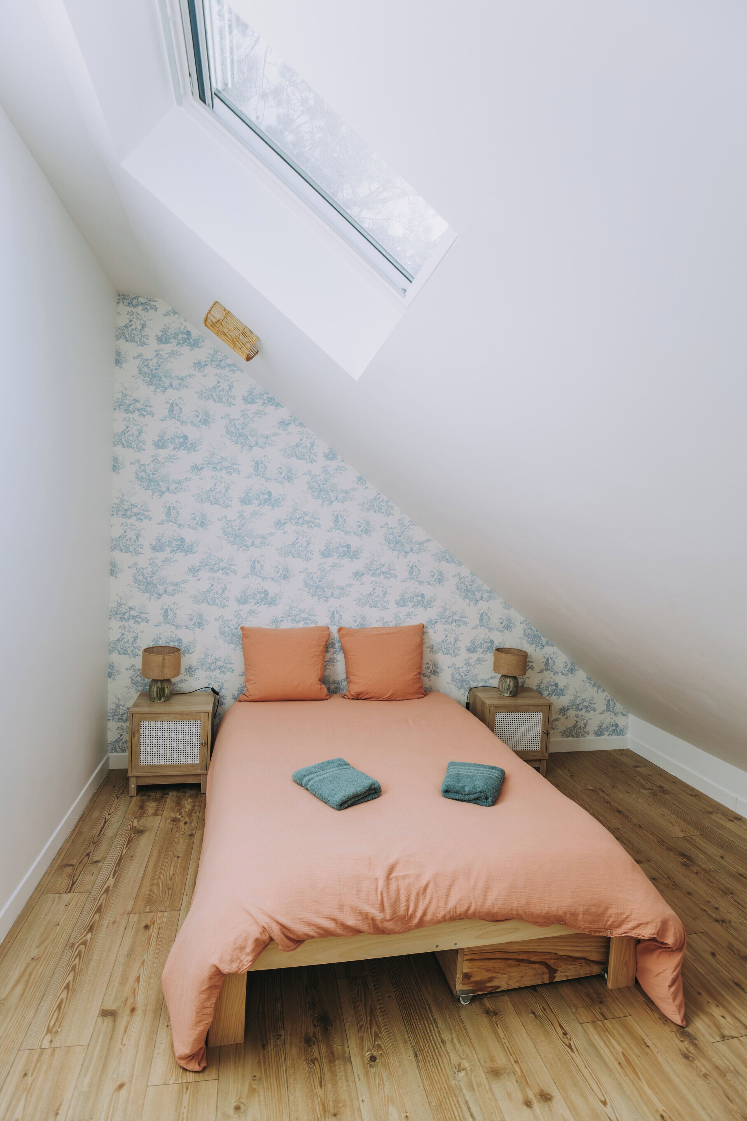 Un dormitorio con techo inclinado, una cama con dos almohadas, dos mesitas de noche con lámparas y un ventanal con vistas a árboles.