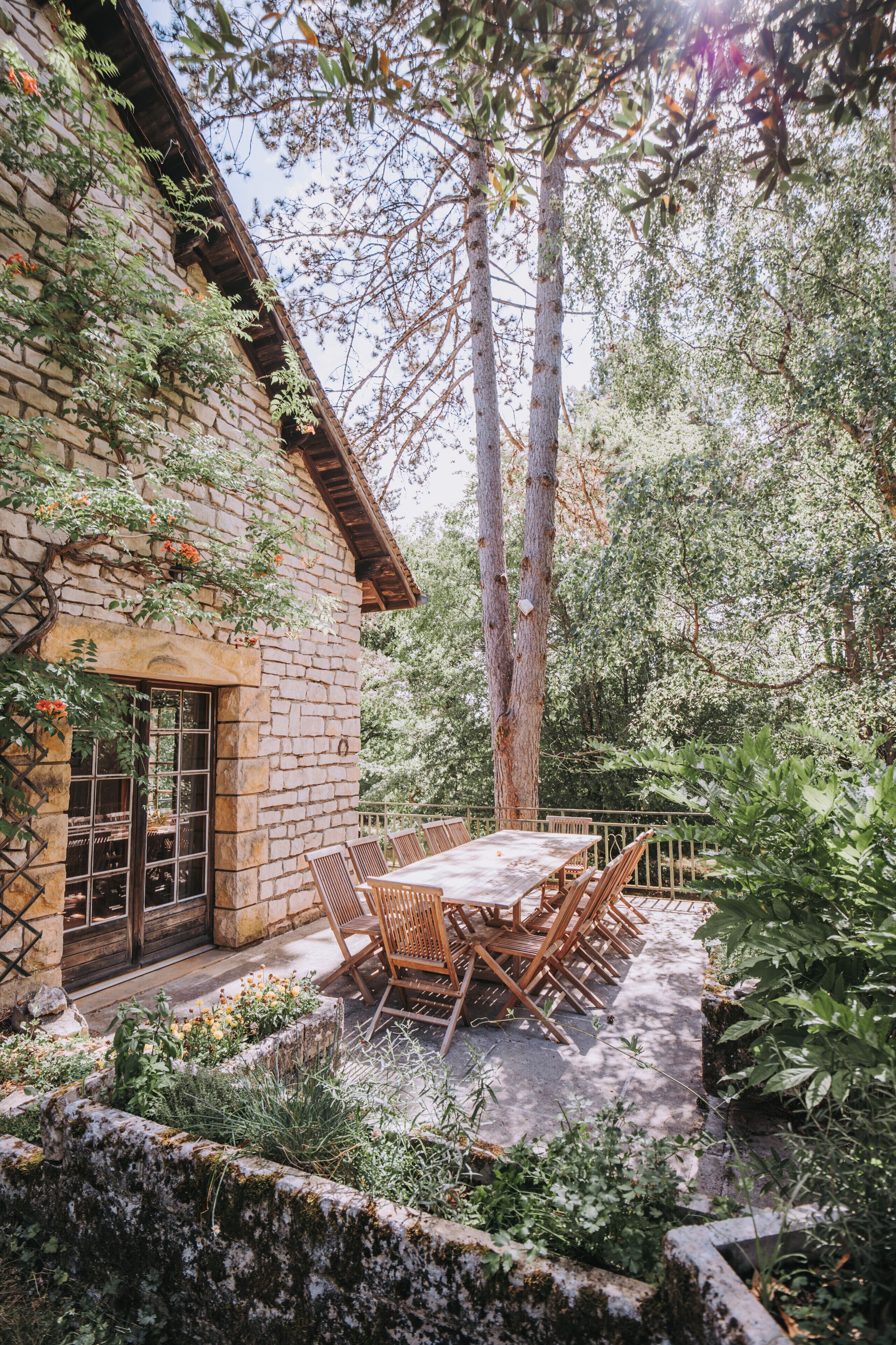 Un bâtiment en pierre avec une table et des chaises en bois, entouré de verdure et d’arbres.