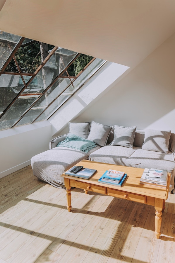 A cozy loft bedroom with a sloped ceiling, a large window, a sofa, and a wooden coffee table with books and remotes.