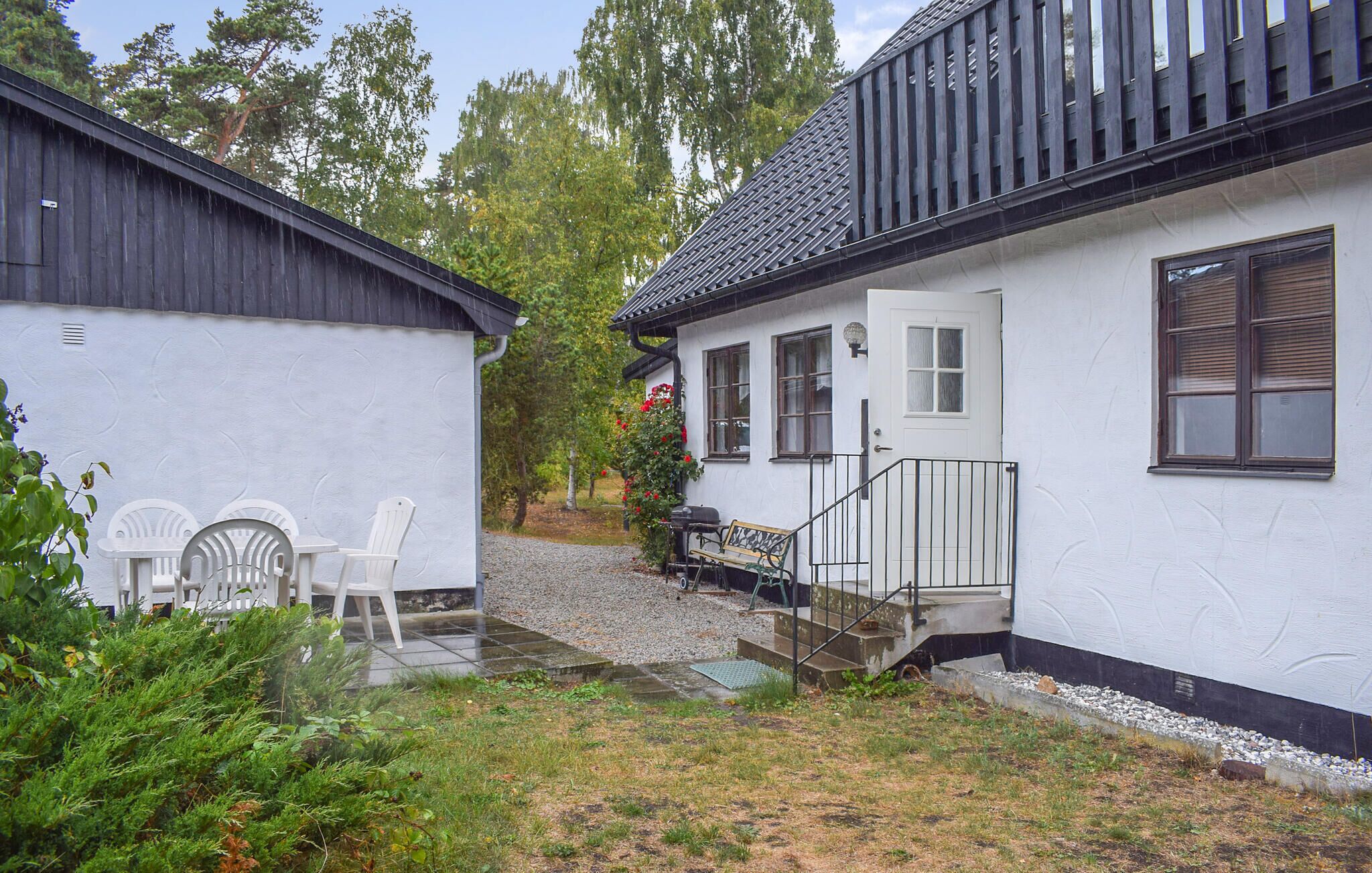A white building with a black roof, a small porch with a bench, and a garden area with chairs and greenery.