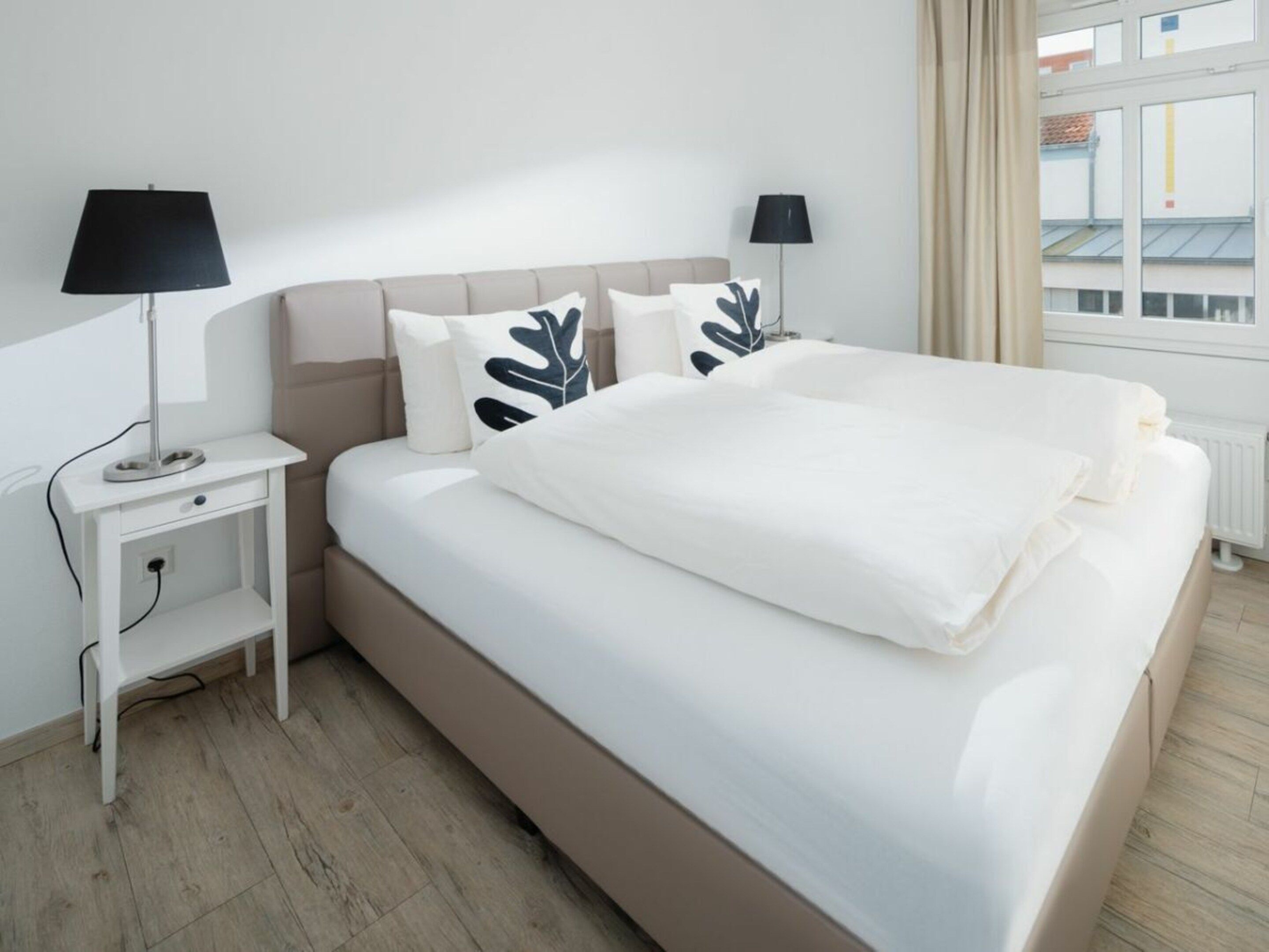 A neatly made bed with a white comforter and pillows, a bedside table with a lamp, and a window with curtains.