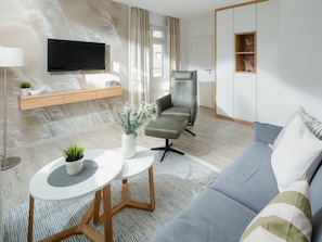 Ein modernes Wohnzimmer mit einer Couch, einem Sessel, einem kleinen Tisch mit einer Pflanze, einem Wand-Fernseher und einer Holzkommode mit Ablagefläche.