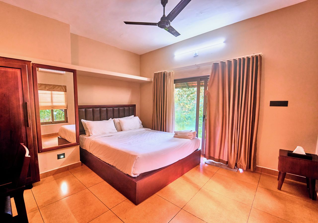 Quarto de hotel com uma cama grande, ventoinha no teto, janela com cortinas e mesa de cabeceira.