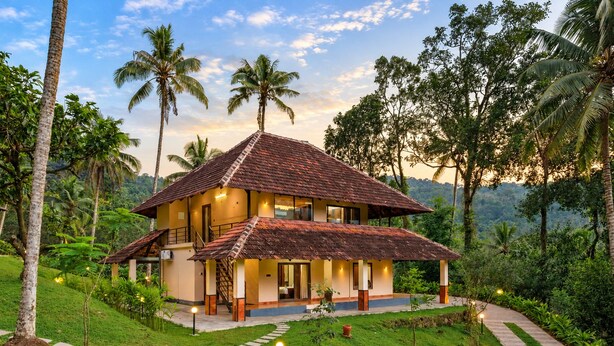 A two-story house with a tiled roof, surrounded by lush greenery and palm trees.