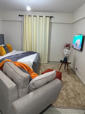 A bedroom with a bed, a TV, a small table with a vase, and a sofa. - Cozy studio apartment in a nice quiet space with a gym and pool (Nairobi)