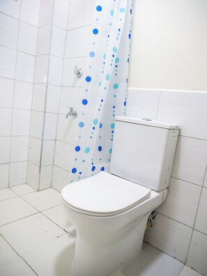 A bathroom with a white toilet, a shower curtain with blue polka dots, and white tiled walls. - Cozy studio apartment in a nice quiet space with a gym and pool (Nairobi)