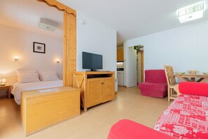 A room with a bed, a wooden cabinet, a television, a red chair, and a dining table. - Appart cosy et charmant, coeur de la station (Les Orres)