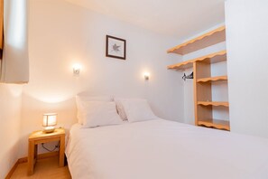 A bedroom with a bed, bedside table, wall-mounted light, and a wooden shelf unit. - Appart cosy et charmant, coeur de la station (Les Orres)