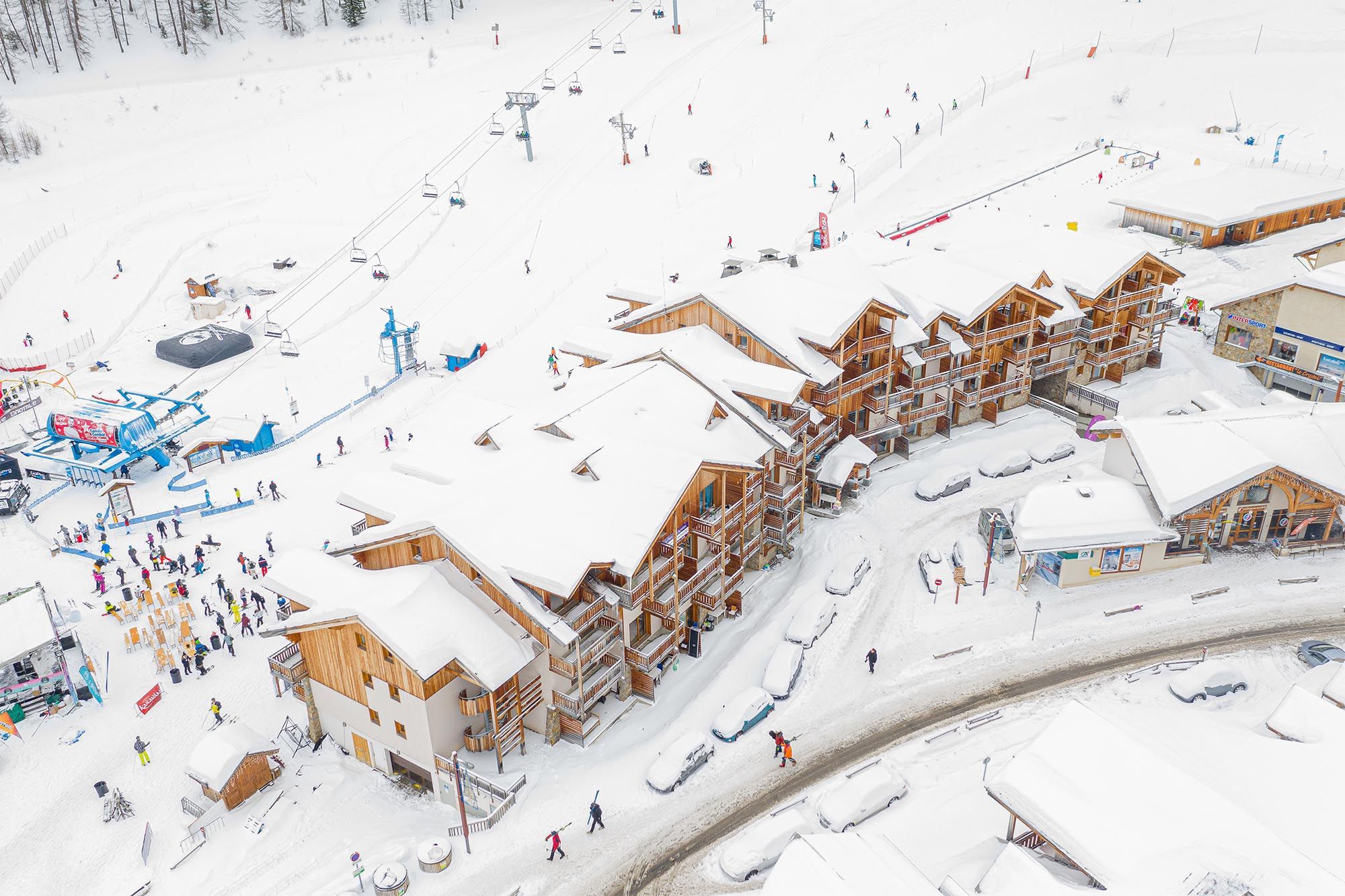 A snow-covered ski resort with wooden buildings, ski lifts, and a parking area.