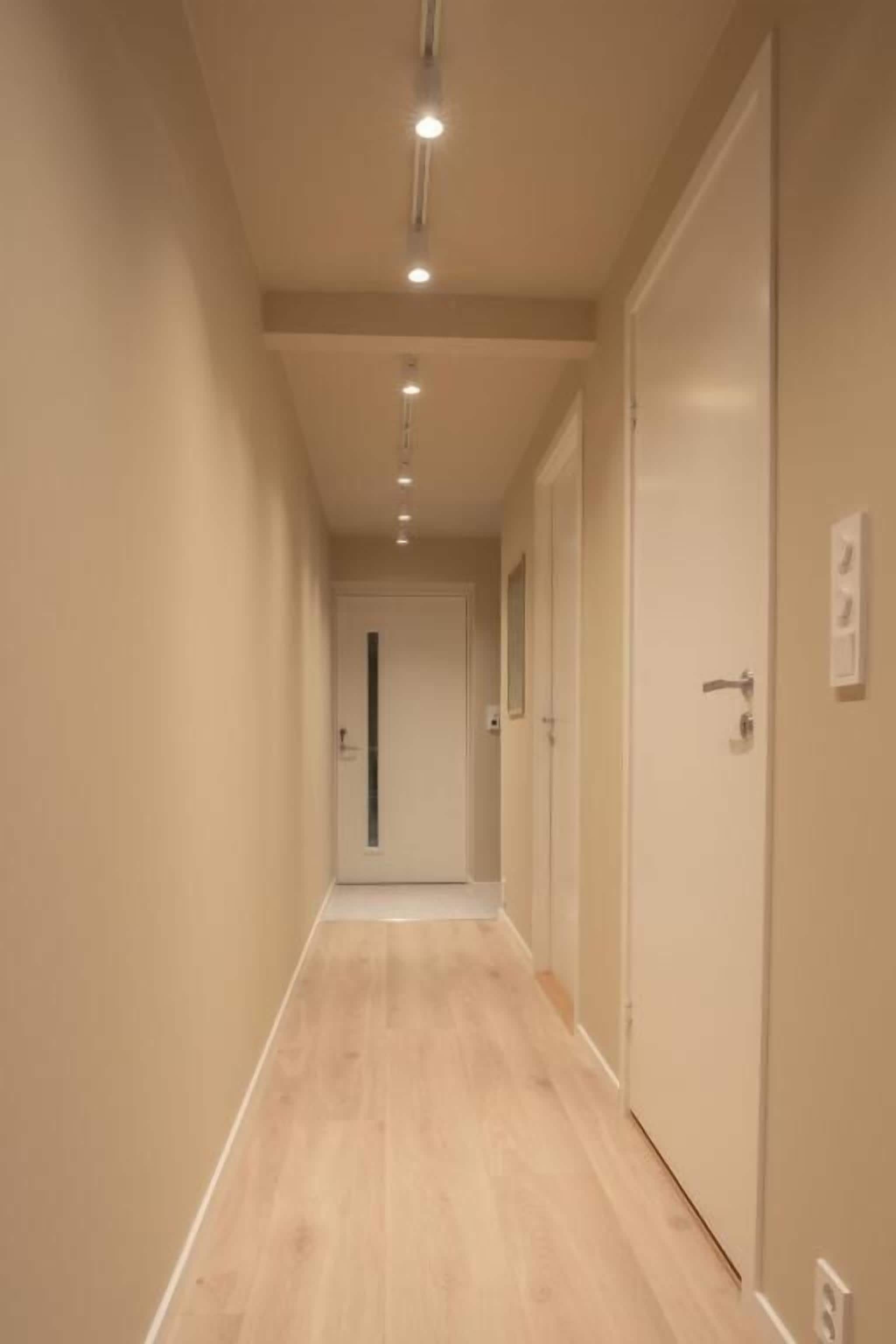Een hal met een houten vloer, deuren aan de rechterkant en inbouwverlichting in het plafond.