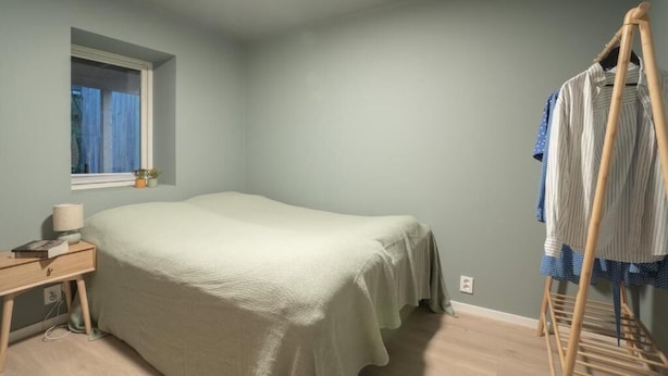 Un dormitorio con una cama individual, una mesita de noche con lámpara, una ventana y un tendedero de madera con ropa.