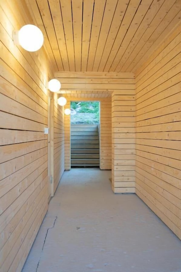 A narrow wooden hallway with a ceiling and walls made of horizontal wooden planks. There are three spherical light fixtures on the left wall.