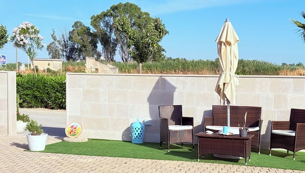 An outdoor seating area with wicker furniture and a beige umbrella, set against a stone wall with greenery in the background.