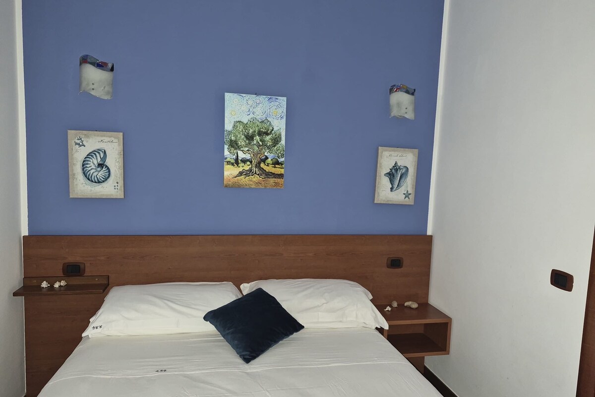 A bedroom with a wooden headboard, a bed with white bedding and a dark blue pillow, and four wall-mounted artworks depicting shells and a tree.