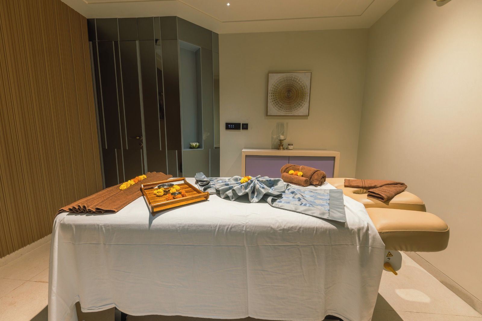 A massage room with a bed, towels, a tray with flowers, and a wall-mounted artwork.