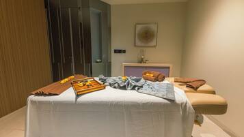 A massage room with a bed, towels, a tray with flowers, and a wall-mounted artwork.