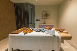 A massage room with a bed, towels, a tray with flowers, and a wall-mounted artwork.