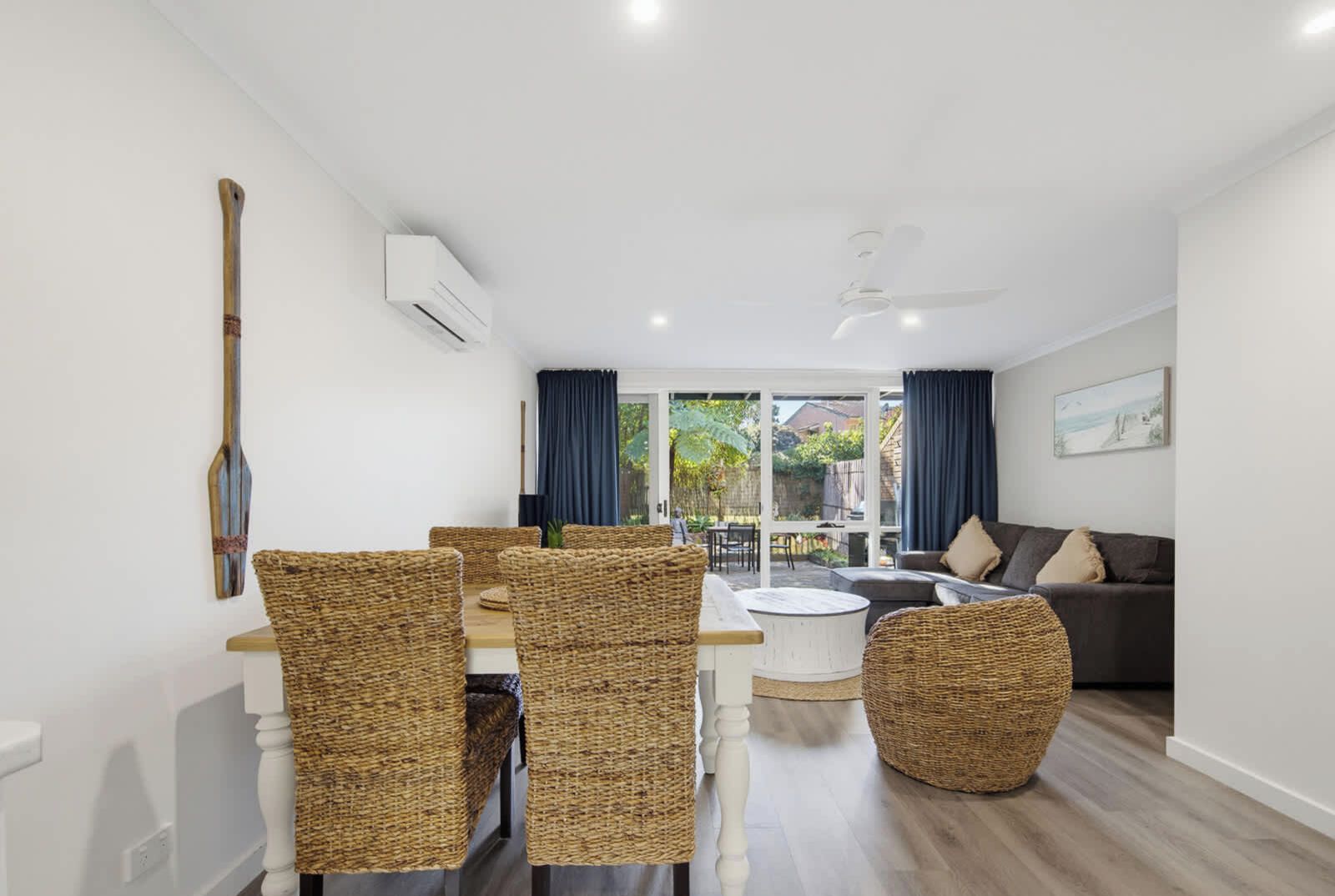 A modern living room with a dining area, featuring wicker furniture, a ceiling fan, and a view through glass doors.