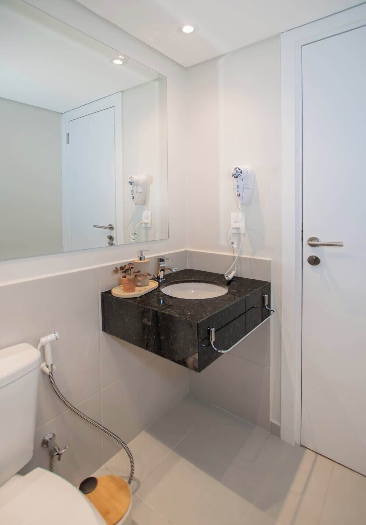 Une salle de bain moderne avec des toilettes blanches, un lavabo noir doté d’un plan de travail en marbre, un sèche-cheveux et une douche avec un flexible.