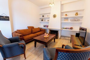 Uma sala de estar com sofá, poltronas, mesa de centro, lareira, estantes com livros e objetos decorativos, e uma televisão.