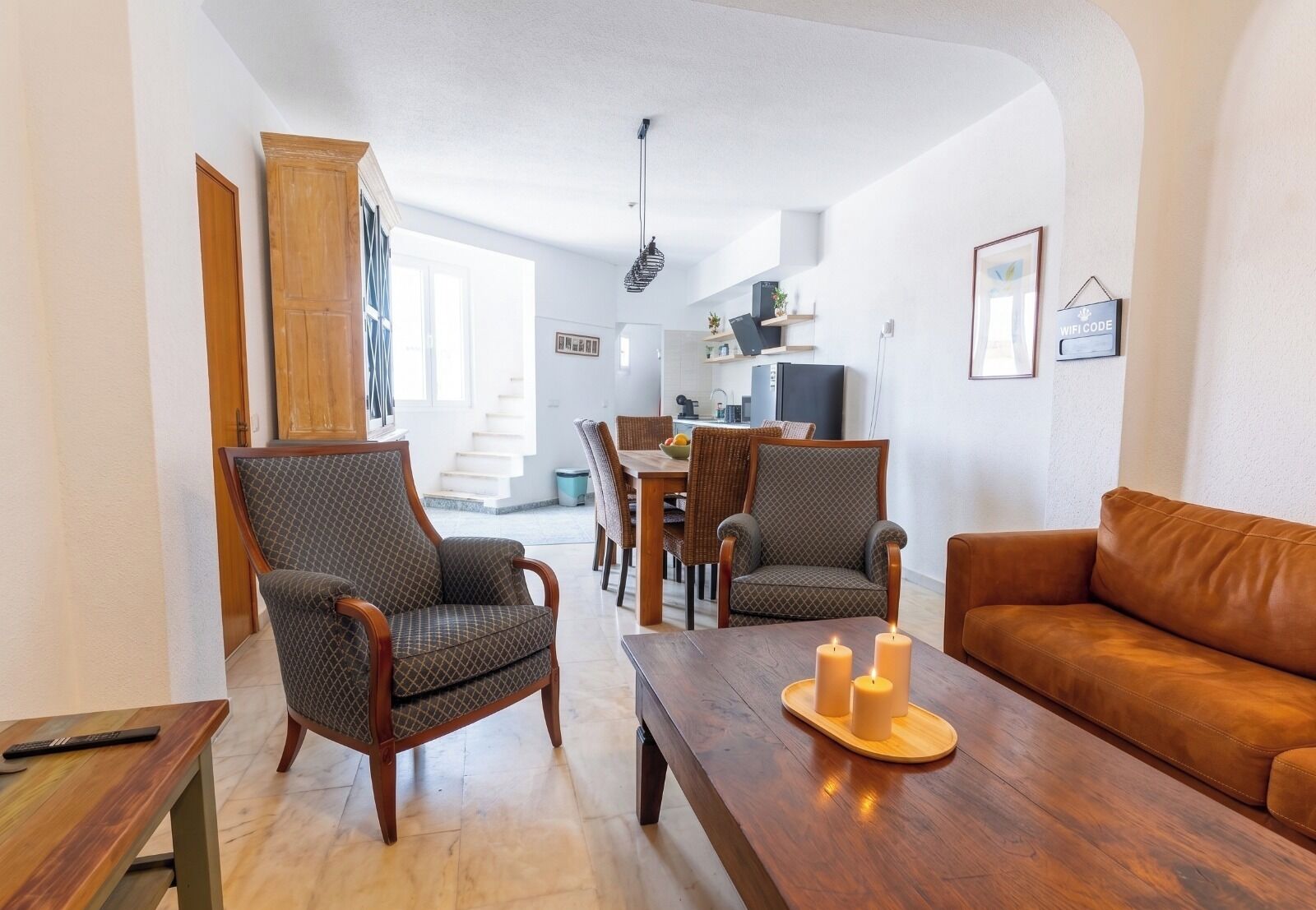 A living room with a brown leather sofa, two patterned armchairs, a wooden coffee table with candles, and a dining area in the background.