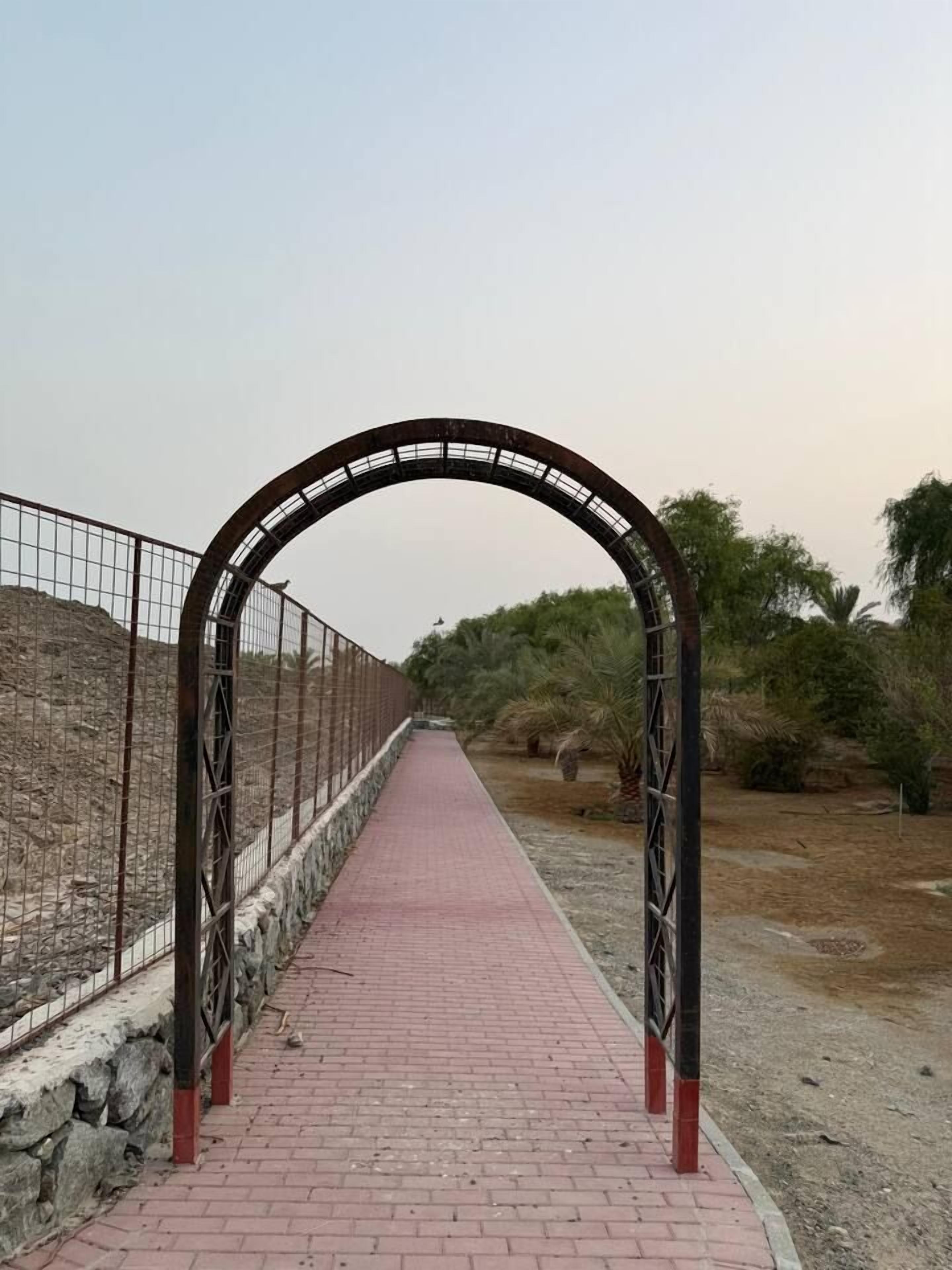 Un sentier bordé d’une arche métallique qui mène à un espace paysager agrémenté d’arbres et d’arbustes.