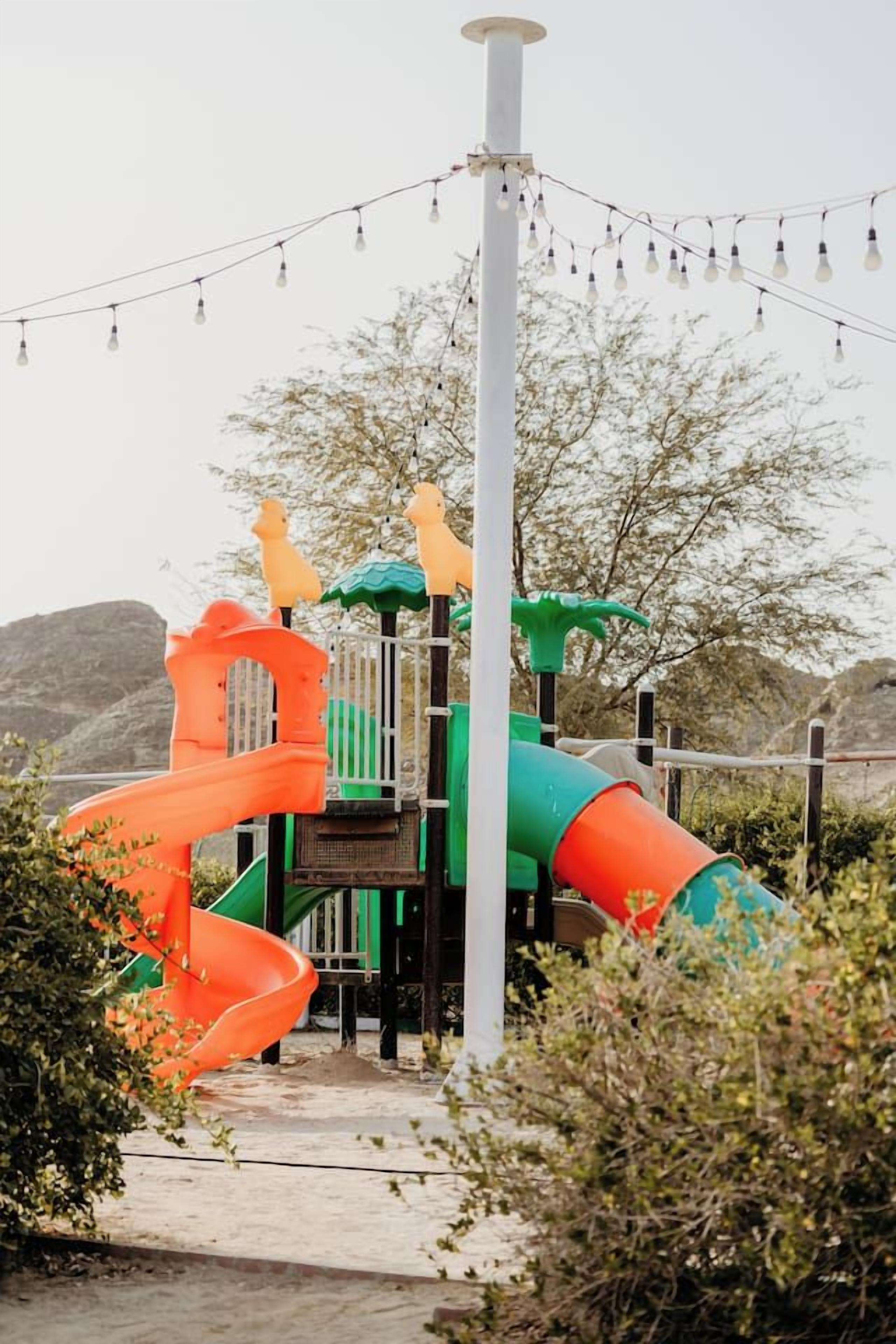 A playground with a slide and climbing structures, surrounded by vegetation and a clear sky.